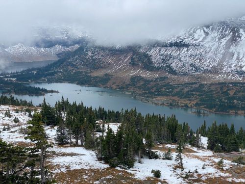 A scenic view of a lake surrounded by mountains and evergreen trees, with patches of snow and cloudy skies.