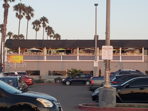 A restaurant with outdoor seating on a second floor, surrounded by palm trees and parked cars in the foreground.