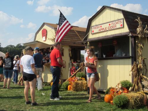 A busy outdoor market scene with people at food stalls, pumpkins, and autumn decorations, featuring an American flag.