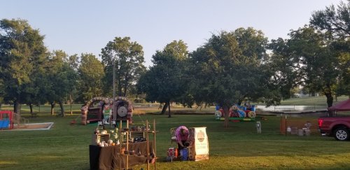 A park scene with inflatable attractions, a vendor stand, and trees, set against a clear sky and calm water.