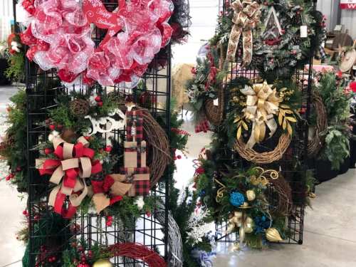 Display of festive holiday wreaths in various styles and colors, arranged on black wire stands.