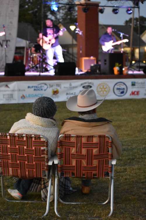 A couple sits in lawn chairs, watching a live band perform on stage at an outdoor event.