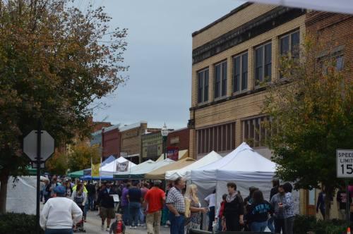 A bustling street market with tents and people, surrounded by brick buildings and autumn trees.
