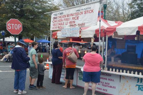 A food stall at a fair with a sign for Thai Town, featuring people ordering and a menu of various dishes.