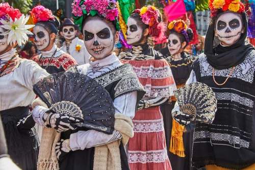 A group of people in traditional attire with skull makeup and colorful flowers, holding fans during a festive celebration.