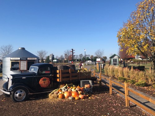 A vintage truck with pumpkins parked near a farm, surrounded by autumn foliage and a clear blue sky.