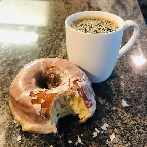 A glazed donut with a bite taken out, next to a white coffee mug filled with black coffee on a granite countertop.