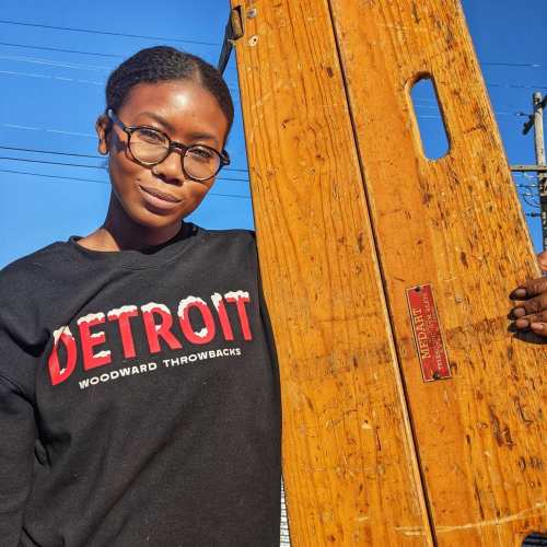 A person wearing a black "Detroit" sweatshirt stands next to a wooden board, smiling in bright sunlight.