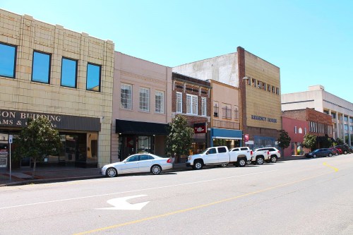 A sunny street view of a small town with various shops and buildings, featuring parked cars along the curb.