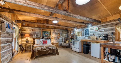 Cozy cabin interior with wooden beams, a bed, kitchenette, and rustic decor, creating a warm, inviting atmosphere.