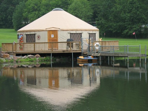 A round yurt on stilts by a calm lake, with a wooden deck and a kayak nearby, surrounded by greenery.