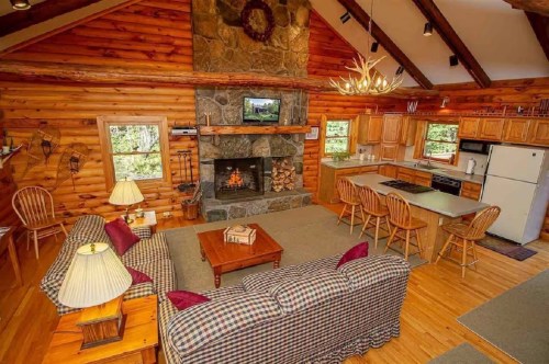 Cozy log cabin interior with a stone fireplace, kitchen, and comfortable seating area. Warm, rustic decor throughout.