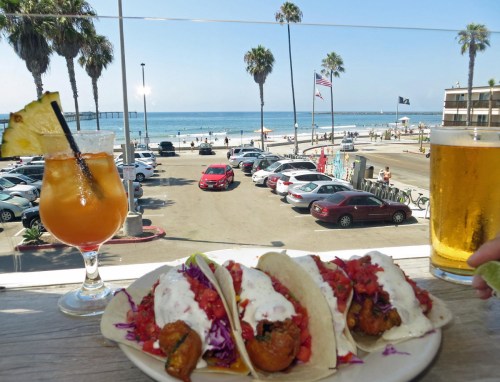 A plate of tacos with shrimp and toppings, a cocktail, and a beer, with a beach view and palm trees in the background.
