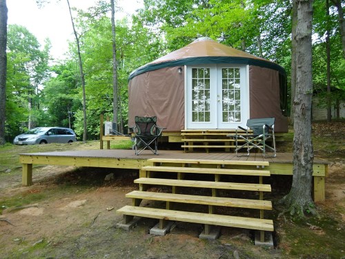 A cozy yurt on a wooden deck surrounded by trees, with two chairs and a car parked nearby.