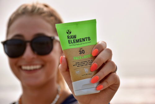 A person wearing sunglasses holds a tube of Raw Elements sunscreen, smiling against a beach backdrop.
