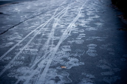 Footprints and tire tracks create patterns on a wet, gray pavement, showcasing a mix of human and vehicle movement.