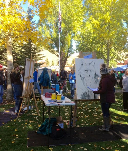 A woman paints an owl on a canvas at an outdoor art event, surrounded by trees and a crowd of onlookers.