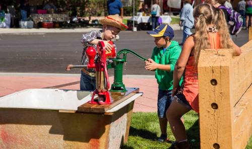 Two children operate hand pumps at a water station, while a girl watches nearby, in a lively outdoor setting.