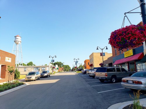 A quiet street lined with shops and parked cars, featuring a water tower in the background and hanging flower baskets.