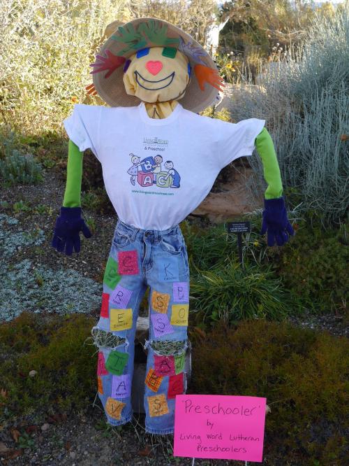 A colorful scarecrow wearing a t-shirt and jeans, decorated with letters and a sign reading "Preschooler."