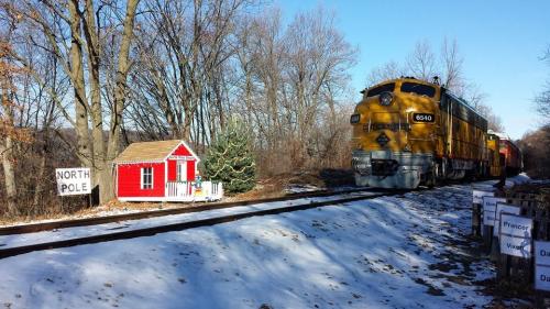 A yellow train passes by a small red cabin labeled "North Pole" amidst snowy trees.