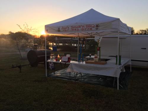 A BBQ tent with a sign reading "Joe's Smokin' BBQ" at sunset, featuring a grill and tables set up nearby.