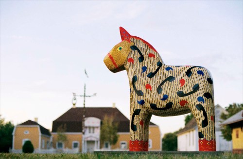 A colorful wooden horse sculpture stands in front of a historic building under a clear sky.