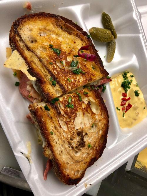 A toasted sandwich with layers of meat and cheese, garnished with herbs, served with pickles and a side of sauce.