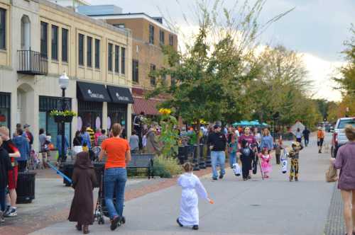 Children in costumes walk along a busy street, with families and shops in the background, celebrating a festive event.