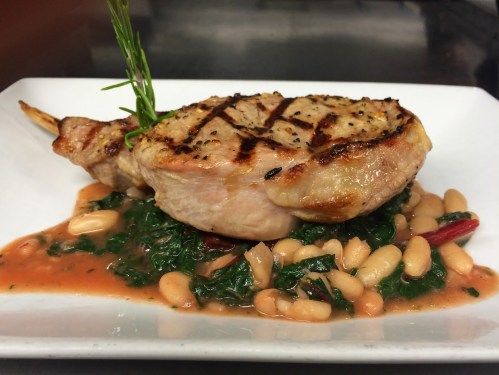 Grilled pork chop on a bed of sautéed greens and white beans, garnished with a sprig of rosemary.