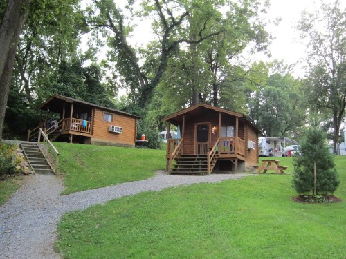 Two wooden cabins on a grassy area, surrounded by trees, with a gravel path and picnic table nearby.