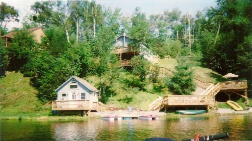 A serene lakeside scene featuring wooden cabins, lush greenery, and a dock with boats on calm water.