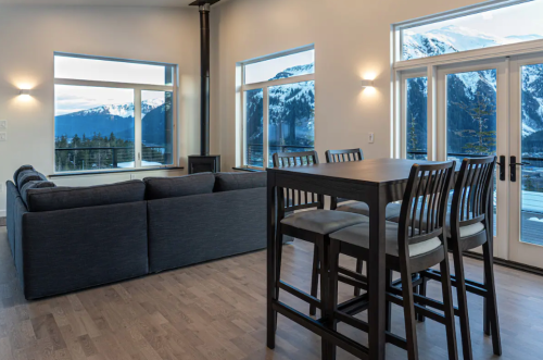 Modern living room with large windows showcasing mountains, featuring a gray sofa and a tall dining table with chairs.
