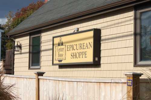 Sign for "Epicurean Shoppe" on a building with a wooden fence in front, under a partly cloudy sky.