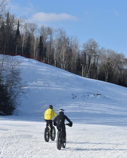 Two cyclists ride fat bikes on a snowy landscape, with trees and a hill in the background under a clear blue sky.