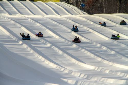 A snowy tubing hill with several people sliding down in colorful inner tubes, surrounded by trees.