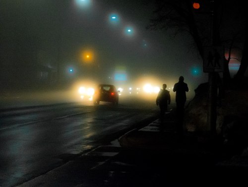 Two silhouettes walk along a foggy street, with blurred headlights illuminating the misty scene.
