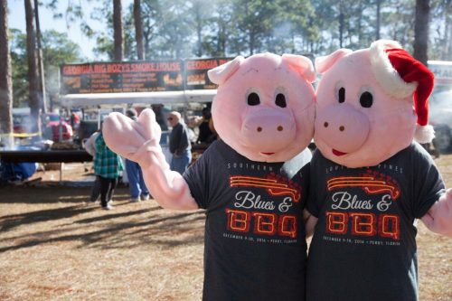 Two people in pig costumes wave at a BBQ festival, with food stalls and trees in the background.