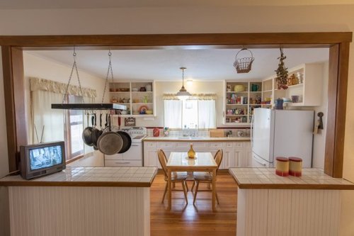 A cozy kitchen with white cabinets, a dining table, and a vintage TV, featuring hanging pots and shelves filled with dishes.