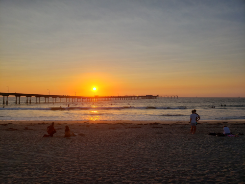 Sunset over a beach with a pier, people sitting on the sand, and surfers in the water.