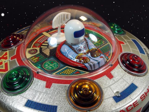 A colorful toy spaceship with a dome, featuring a seated astronaut figure and bright buttons, set against a starry background.