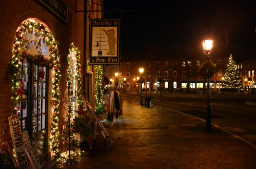 A festive street at night, adorned with holiday lights and decorations, featuring a shop with a sign and a lamppost.