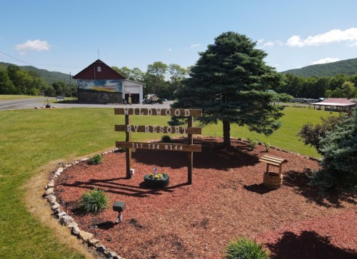 A wooden sign for "Woodwood RV Resort" surrounded by landscaped greenery and a barn in the background.