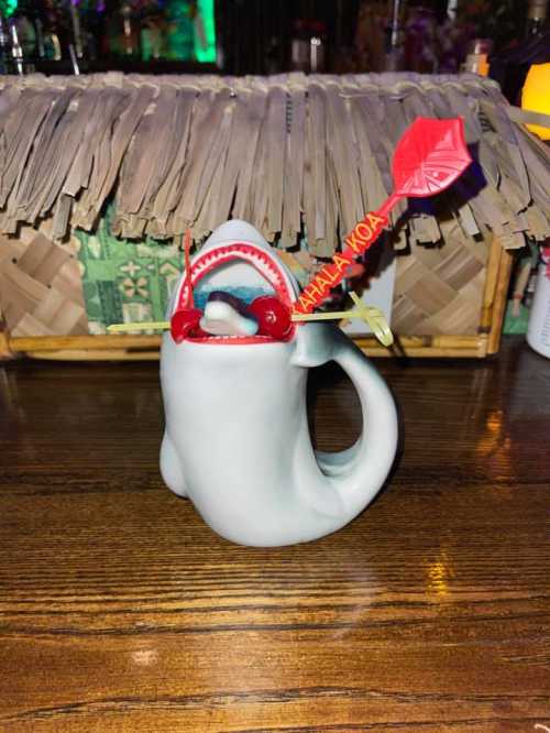 A shark-shaped mug with a red straw and a decorative umbrella, sitting on a wooden table.