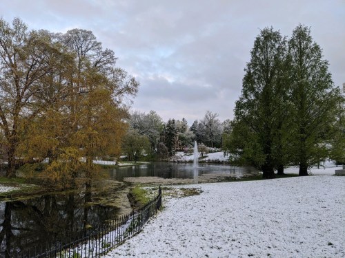 A serene winter scene featuring a pond surrounded by snow-covered trees and a fountain in the background.