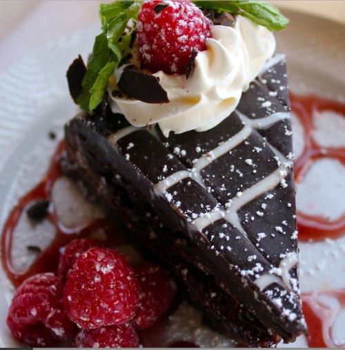 A slice of rich chocolate cake topped with whipped cream, raspberries, and mint, drizzled with sauce on a plate.