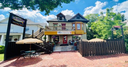A charming two-story house with a colorful facade, surrounded by greenery and featuring a patio with umbrellas.