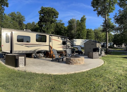 A cozy RV parked on a circular patio surrounded by trees, with a fire pit and a small shed nearby.