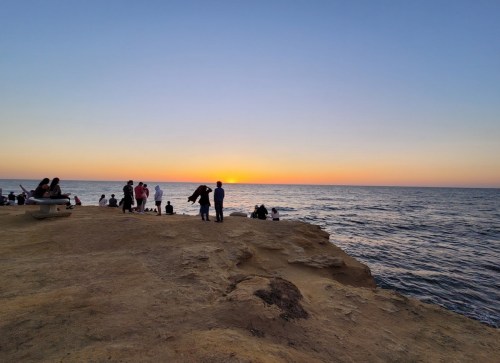 People gather on a rocky shore at sunset, with the sun dipping below the horizon over the calm ocean.