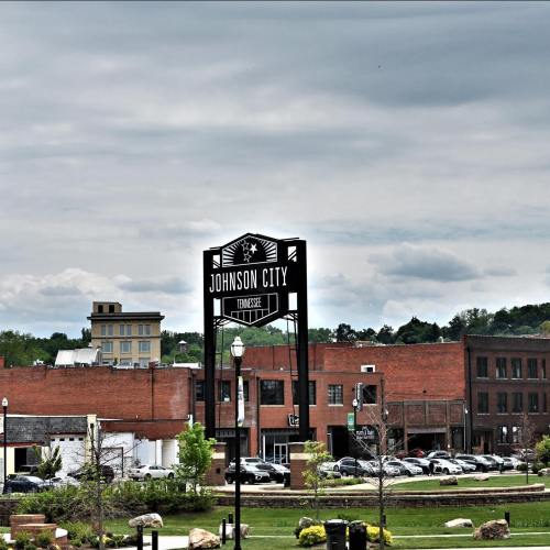Sign for Johnson City, Tennessee, with a backdrop of buildings and a cloudy sky. Greenery in the foreground.